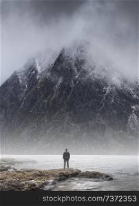 Man stood on lake shore dwarfed by mountain in background concept image of challenge and achievement