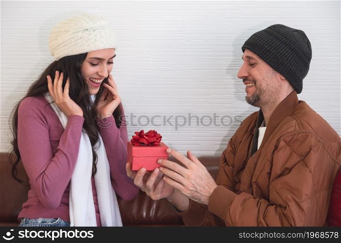 Man surprising his girlfriend with red gift box on couch in living room. Couple celebrating holidays, anniversary or birthday together at home.