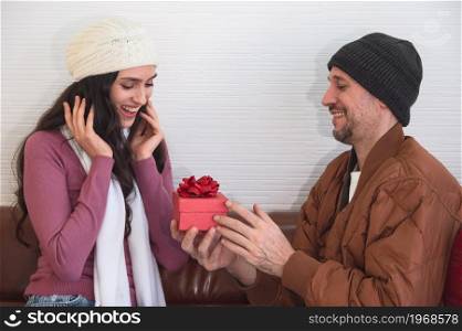 Man surprising his girlfriend with red gift box on couch in living room. Couple celebrating holidays, anniversary or birthday together at home.