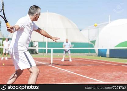 Man swinging tennis racket while playing doubles match on red court during summer weekend