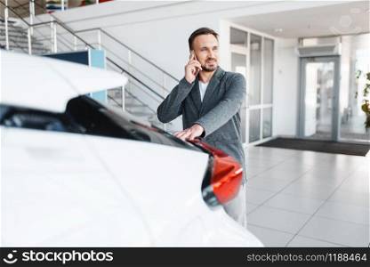 Man talking by phone near the new car in showroom. Male customer buying vehicle in dealership, automobile sale, auto purchase. Man talking by phone near the new car in showroom