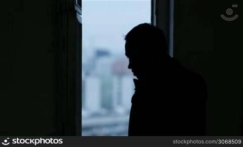 Man talking on the phone near the window. Black silhouette on blurry city background in the window.