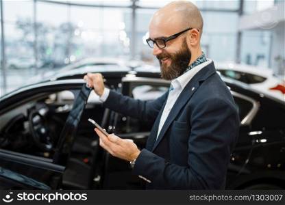 Man talks by mobile phone near new automobile in car dealership. Customer in vehicle showroom, male person buying transport, auto dealer business. Man talks by mobile phone in car dealership