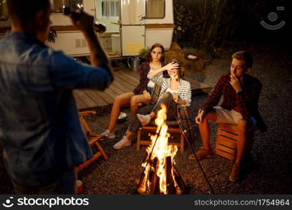 Man tells a scary story to his friends by the bonfire, picnic at camping in the forest. Youth having summer adventure on rv, camping-car on background. Two couples leisures, travelling with trailer. Man tells a scary story by the bonfire, camping