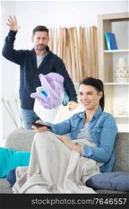 man throwing laundry on lazy wife sitting on sofa
