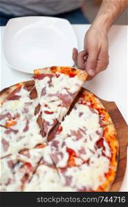 Man took first slice of delicious Italian pizza. In frame hand taking slice of hot pizza with ham on wooden board, white serving plate on white table. Pizza time. Top view