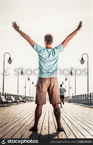 Man tourist on pier with raised hands arms. Guy enjoying summer travel vacation by sea. Fashion. Happiness and freedom concept.