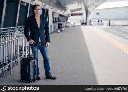 Man traveler poses with suitcase on wheels wears protective mask during pandemic outbreak afraids of dangerous influenza coronavirus posses at platform waits for transport. Health, safety life