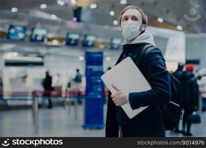 Man traveler wears protective disposable medical mask in airport, returns from abroad where coronavirus spreading, carries backpack, takes care of health, protects from virus, has postponed flight