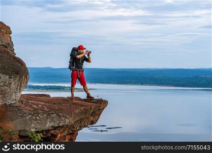man traveler with backpack using camera taking a photo on the edge of cliff, on a top of the rock mountain