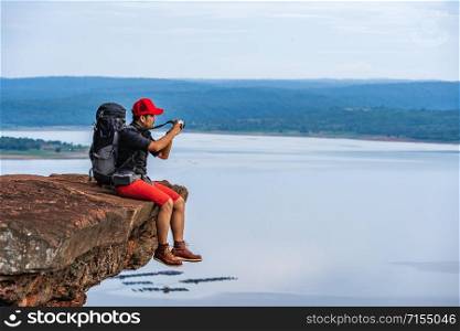 man traveler with backpack using camera taking a photo on the edge of cliff, on a top of the rock mountain