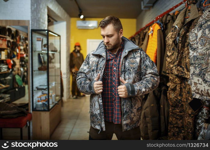 Man trying on uniform at showcase in gun shop. Euqipment and rifles for hunters on stand in weapon store, hunting and sport shooting hobby. Man trying on uniform at showcase in gun shop