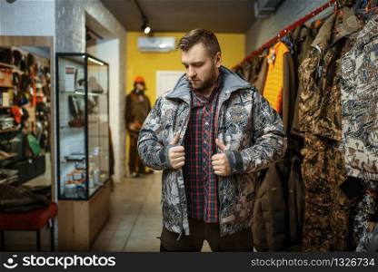 Man trying on uniform at showcase in gun shop. Euqipment and rifles for hunters on stand in weapon store, hunting and sport shooting hobby. Man trying on uniform at showcase in gun shop