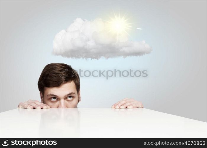 Man under table. Young businessman looking from under table at sun behind cloud