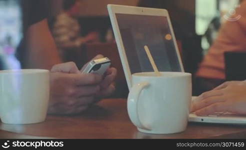 Man uses smartphohe, woman works with laptop while drinking coffee in cafe, faces are invisible