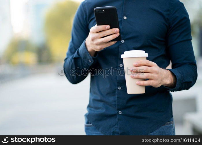 Man using his mobile phone and holding a cup of coffee while standing outdoors at the street. Urban concept.