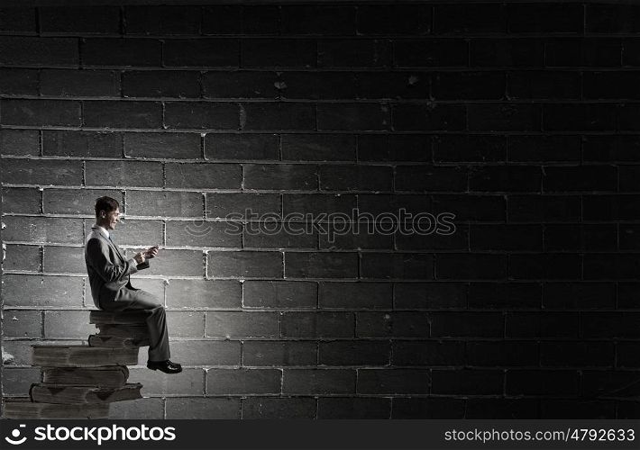 Man using his mobile phone. Young businessman sitting on pile of books with mobile phone in hand