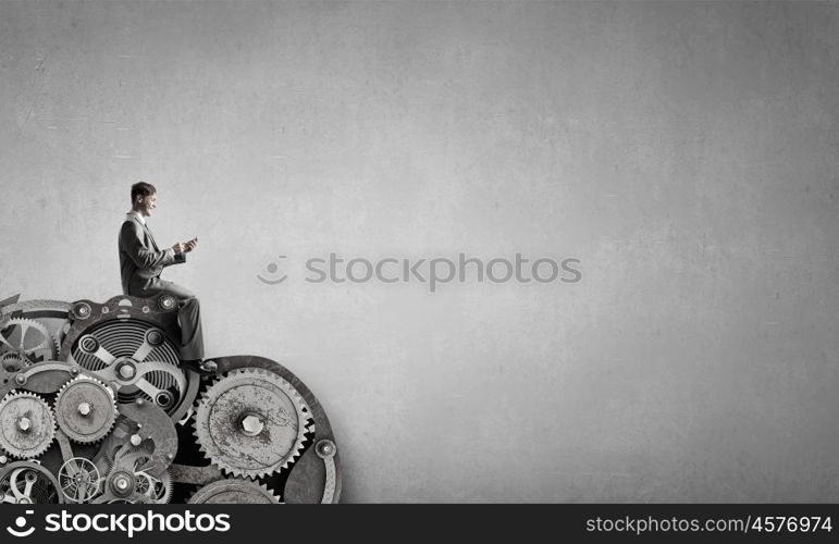 Man using his smartphone application. Young businessman sitting on gears mechanism with mobile phone in hand