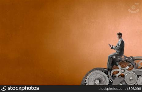 Man using his smartphone application. Young businessman sitting on gears mechanism with mobile phone in hand