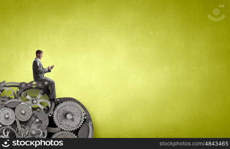Man using his smartphone application. Young businessman sitting on gears mechanism with mobile phone in hand
