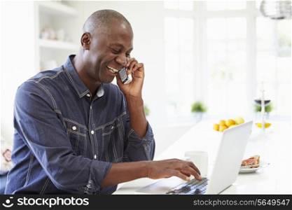Man Using Laptop And Talking On Phone In Kitchen At Home