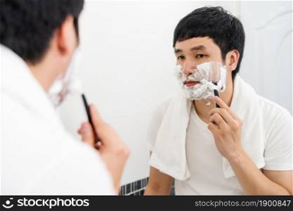 man using razor to shaving his face with cream foam in the bathroom mirror