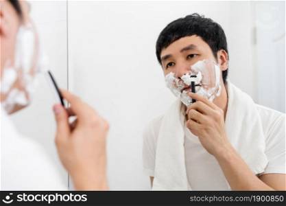 man using razor to shaving his face with cream foam in the bathroom mirror