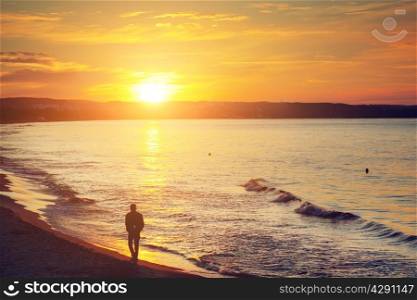 Man walking alone on the beach at sunset. Calm sea with rippling waves.