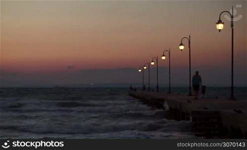 Man walking along the pier with lanterns, some people standing in the distance. Strong sea waves striking pier