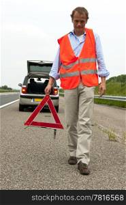 Man walking backwards from his car wearing a safety vest, carying a warning triangle