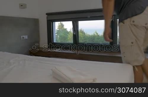Man walking in room and sitting on bed. Using tablet. View on sea from window.