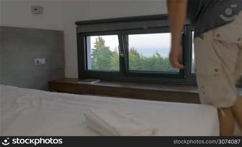Man walking in room and sitting on bed. Using tablet. View on sea from window.