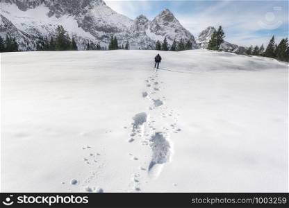 Man walking through snow layer, towards snowy mountain peaks and fir trees. Footsteps in snow. Foot traces in the snow. Winter landscape.