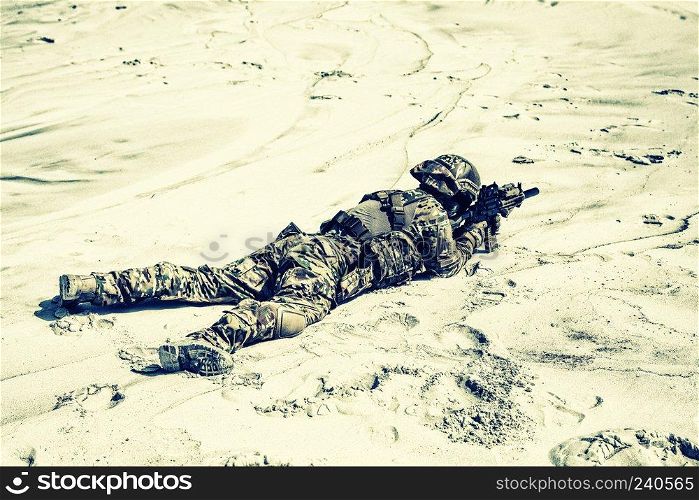 Man wearing camo uniform, equipped with military ammunition, lying on sand and shooting with service rifle replica while playing war game, taking part in desert battle simulation during airsoft event. Airsoft player with rifle in desert skirmish
