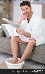 man wearing dressing-gown has feet in footbath and reads newspaper