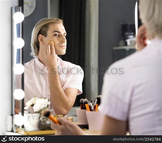 man wearing make up using foundation looking into mirror