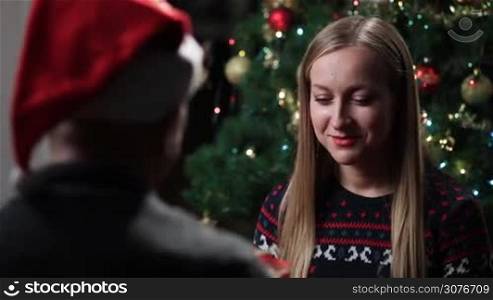 Man wearing Santa&acute;s hat giving christmas gift box to his charming blonde woman and kissing her with tenderness against Christmas tree background. Young couple in love celebrating Christmas at home.