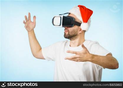 Man wearing santa claus hat exploring space with virtual reality goggles headset, vr box. New generation cyber christmas concept, on blue. Man in virtual reality goggles and santa hat