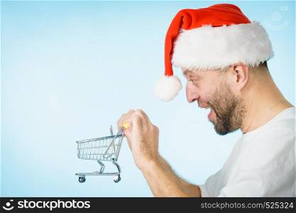 Man wearing santa claus hat holding empty shopping basket cart, on blue. Xmas, seasonal sales, boxing day concept.. Man in santa hat holds empty shopping cart