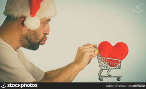 Man wearing santa claus hat holding shopping basket cart with red heart, side view on blue. Christmas, charity sharing concept.. Man in santa hat holds shopping cart with heart.