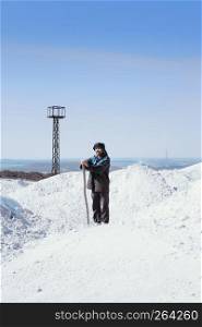 Man with a staff in ethnic clothes standing among mounds of white sand against the background of a distant industrial landscape with a tower under a blue sky. Mountain of extracted gypsum. Moscow region, Russia.
