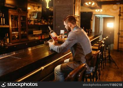 Man with bottle of alcohol beverage drinks at wooden bar counter. Customer leisures in pub, male person relax in restaurant