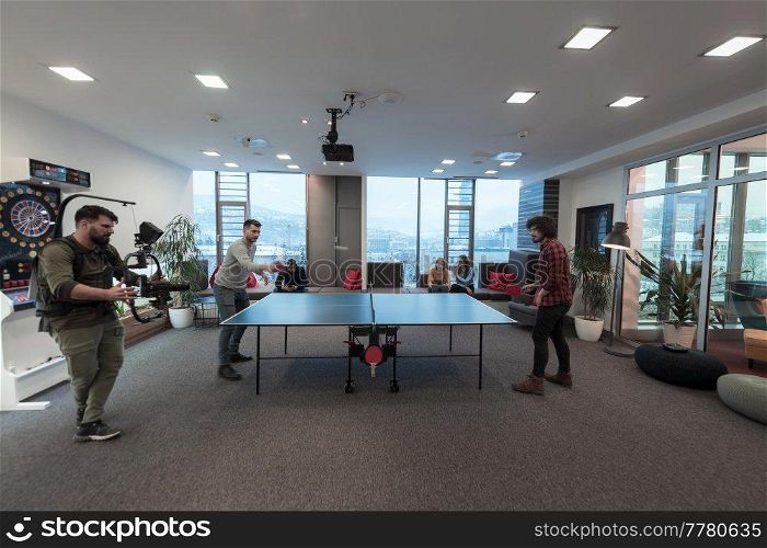 Man with camera recording two young business man playing ping pong tennis at modern creative office space people group have a meeting and brainstorming in background. High-quality photo. Man with camera recording two young business man playing ping pong tennis at modern creative office space people group have meeting and brainstorming in background