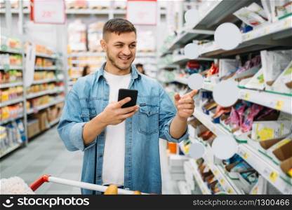 Man with cart makes a purchase by the list on phone in supermarket. Male customer in shop, husband with trolley choosing consumer goods, family shopping