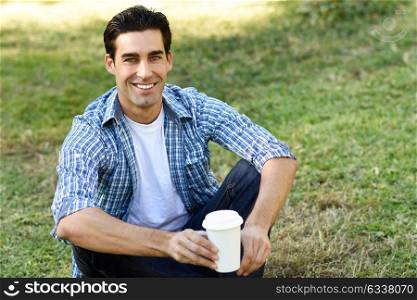 Man with casual clothes drinking coffee to go in an urban park. Man wearing blue plaid shirt and jeans.