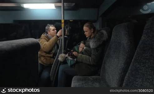 Man with cell taking video of woman with son playing on smart phone while traveling by bus in the evening