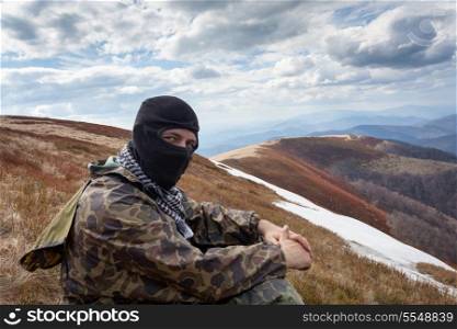 man with closed face and camouflage clothing sitting on a mountain