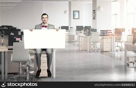 Man with empty banner. Young businessman sitting on table holding white blank banner in modern office