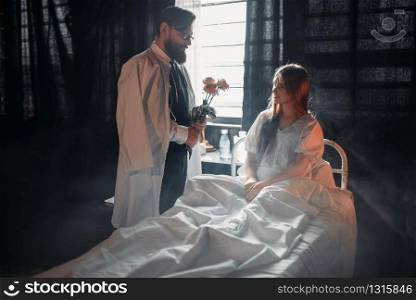 Man with flowers bouquet standing against ill loved woman in hospital bed. Illness of female patient in clinic room, health recovery and treatment. Man with flowers against ill woman in hospital bed