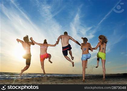 Man with four women running and jumping on beach, back view